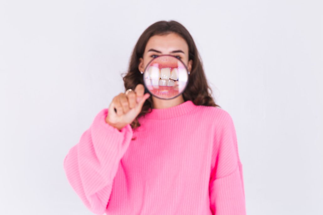 young beautiful woman with freckles light makeup in sweater on white wall with magnifier shows white teeth perfect smile 2